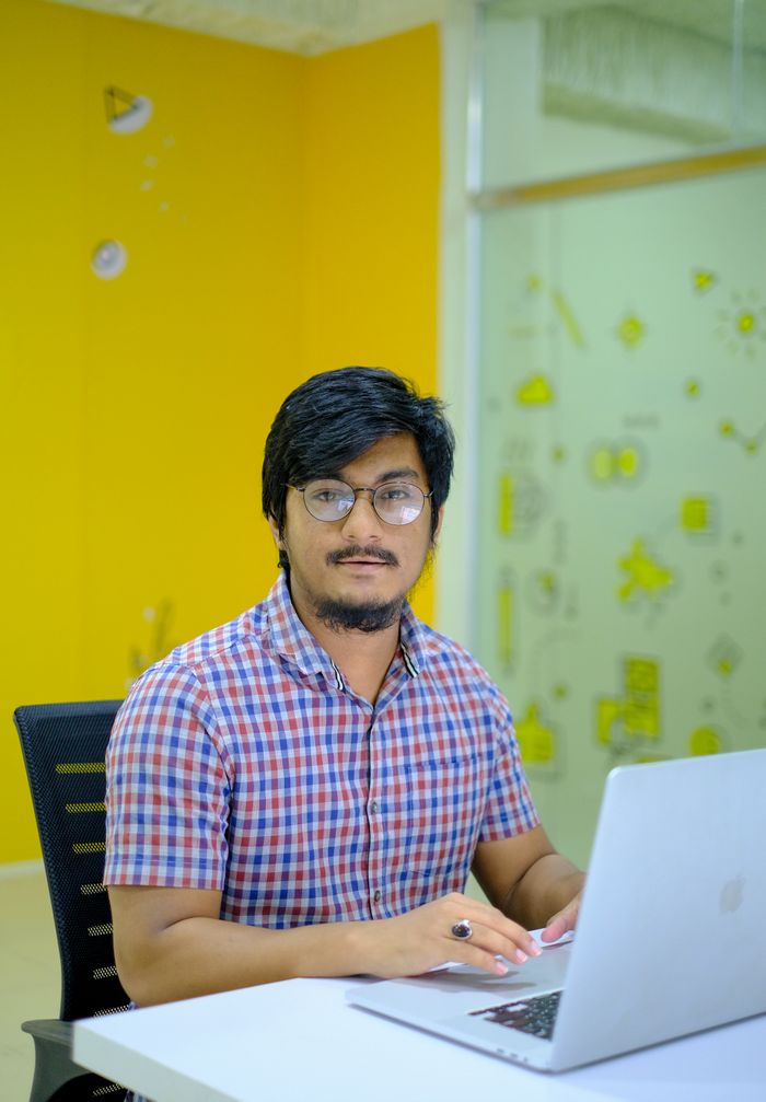 The Art of Drawing Readers In: Your attractive post title goes here Portrait of a young man in an office in Sylhet, Bangladesh, working on a laptop with a modern yellow background.